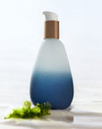 Blue bottle of Mwani Zanzibar’s Face & Body Skin Superfood on sand and next to a piece of seaweed.