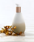 Golden bottle of Mwani Zanzibar’s Facial Cleanser & Mask on sand and next to a piece of seaweed.