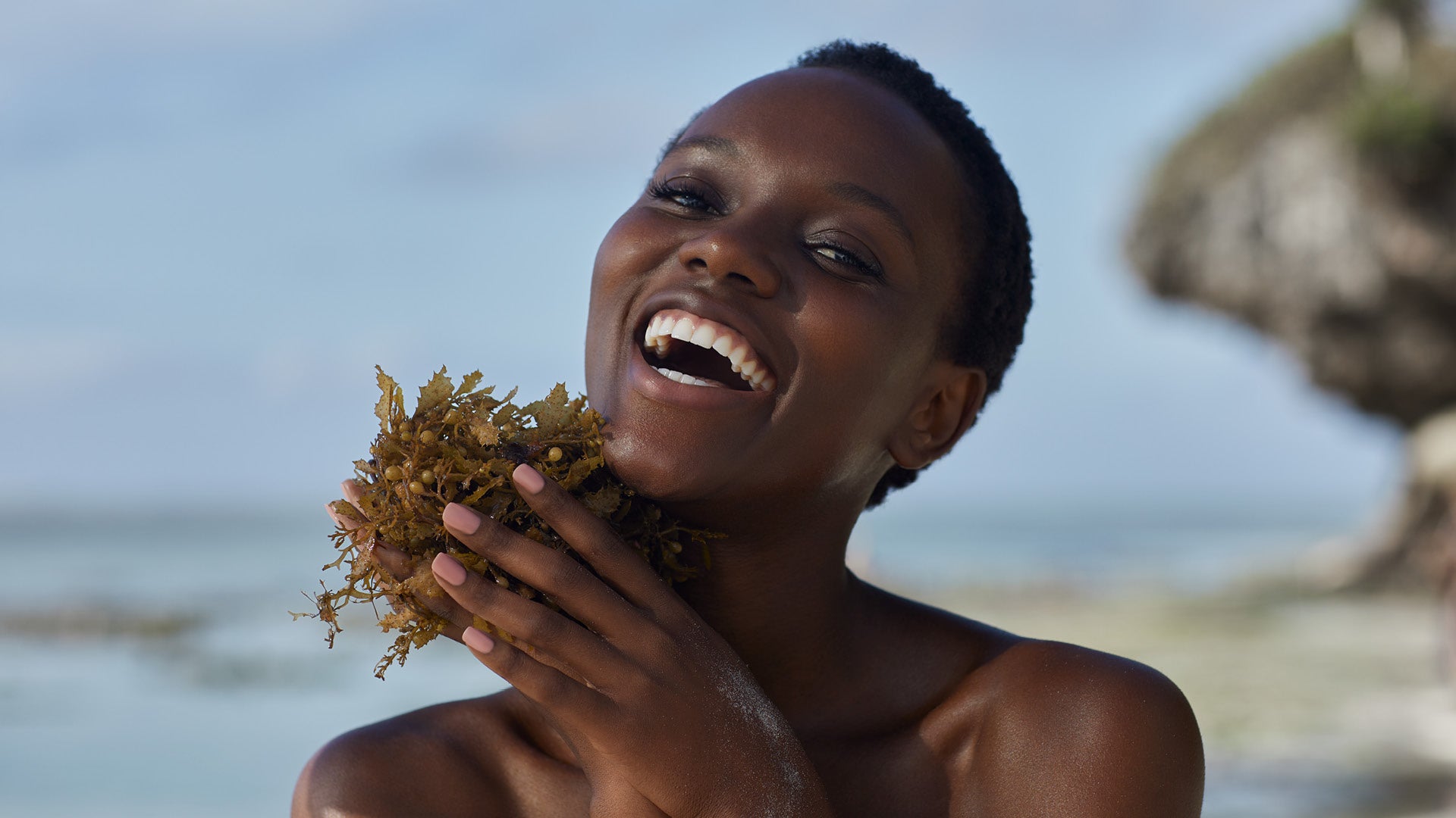 Close up view of Mwani Zanzibar Brand Ambassador Herieth Paul holding seaweed in hand while smiling on an ocean shore in daytime.