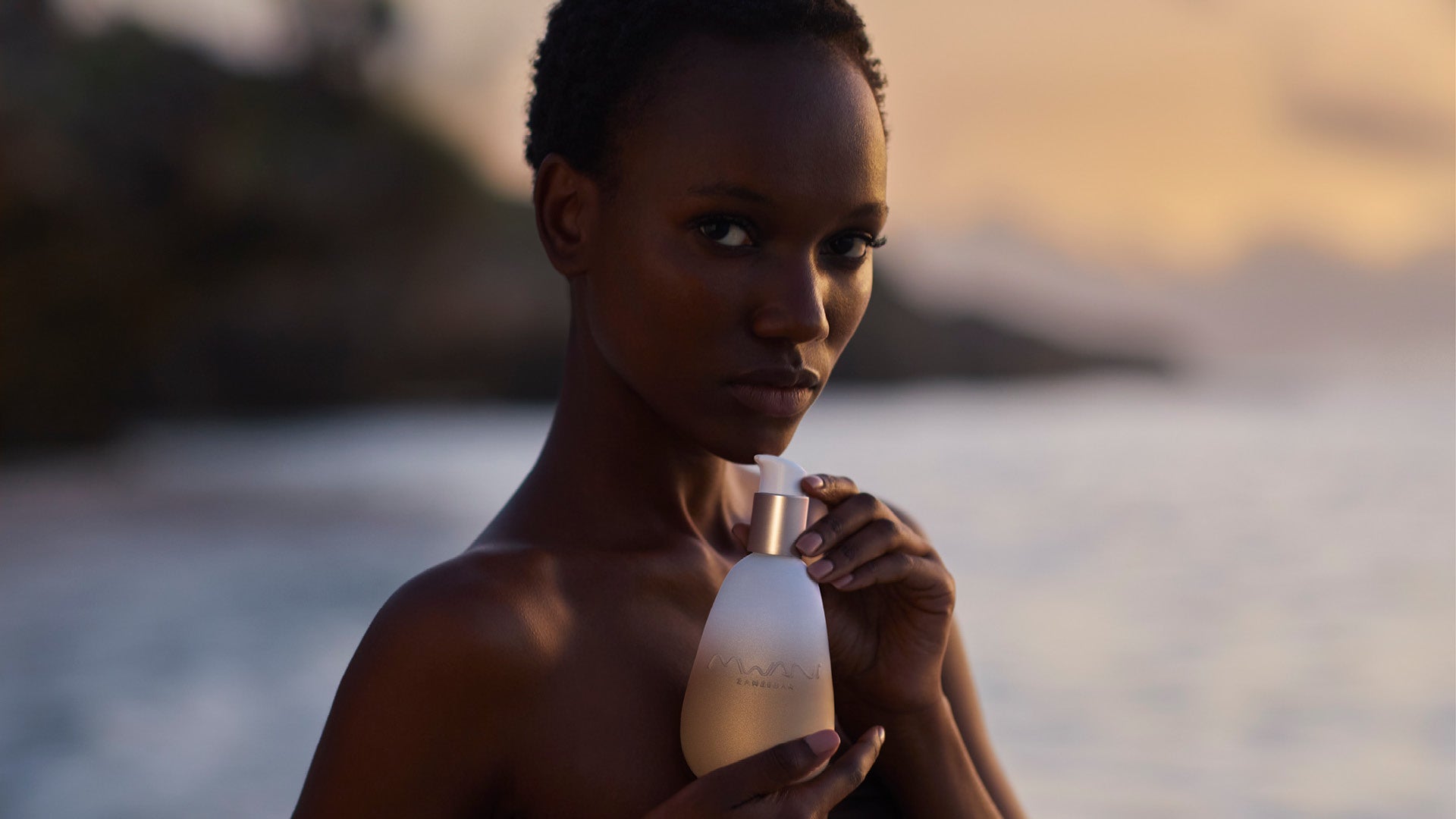 Mwani Zanzibar Brand Ambassador Herieth Paul holding a golden bottle of Mwani Zanzibar’s Facial Cleanser & Mask at dawn in the ocean.