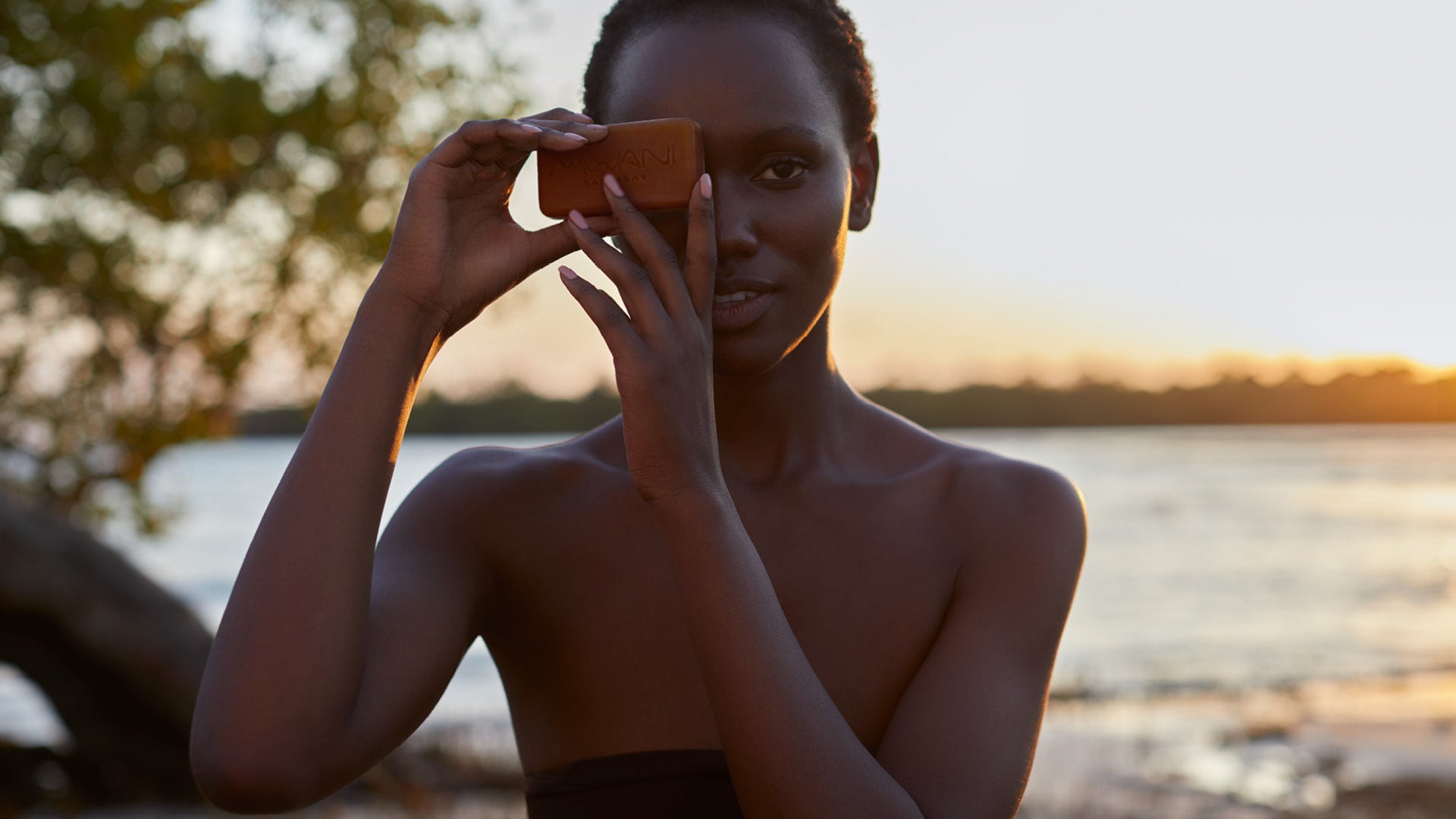 Mwani Zanzibar Brand Ambassador Herieth Paul holding a bar of Mwani Zanzibar’s Seaweed, Cinnamon & Turmeric Soap , standing in front of mangroves and the ocean in the background.